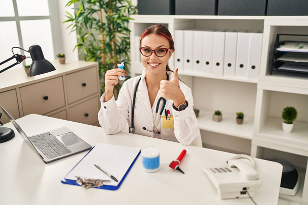 Молода біла жінка в уніформі лікаря тримає бавовняні бруньки посміхаючись щасливим і позитивним, великий палець робить відмінний знак схвалення
  - Фото, зображення