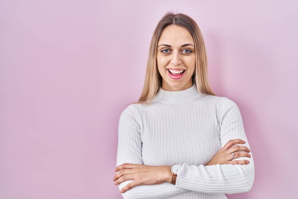 Jeune femme blonde portant un pull blanc sur fond rose heureux visage souriant avec les bras croisés regardant la caméra. personne positive.  - Photo, image