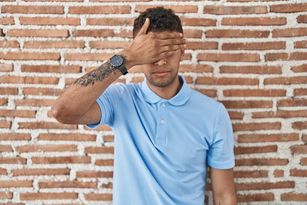 Braziliaanse jongeman die over bakstenen muur staat en ogen bedekt met de hand, en er ernstig en verdrietig uitziet. gezichtsloosheid, verstoppertje en afwijzing  - Foto, afbeelding