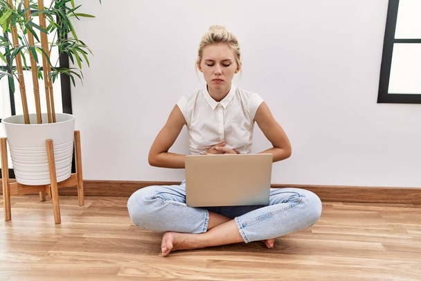 Mulher loira jovem usando laptop computador sentado no chão na sala de estar com a mão no estômago, porque náuseas, doença dolorosa se sentindo mal. conceito de dor.  - Foto, Imagem