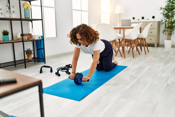 Mujer hispana madura haciendo ejercicio con rueda abdominal en la sala de estar en casa - Foto, imagen