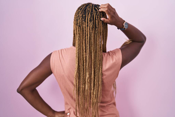 Африканская американка с плетеными волосами, стоящая на розовом фоне задом наперёд, думает о сомнении с рукой на голове  - Фото, изображение