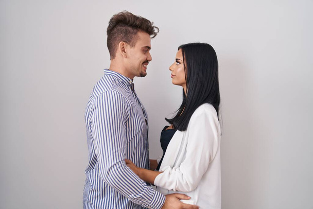 Jeune couple hispanique debout sur fond blanc regardant vers le côté, pose de profil relax avec visage naturel et sourire confiant.  - Photo, image