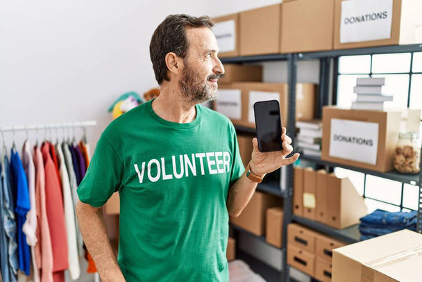Hombre de mediana edad con barba con camiseta voluntaria que sostiene el teléfono inteligente mirando al costado, pose de perfil relajado con rostro natural y sonrisa confiada.  - Foto, imagen