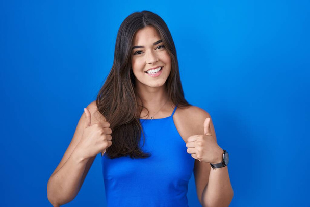 Hispanic woman standing over blue background success sign doing positive gesture with hand, thumbs up smiling and happy. cheerful expression and winner gesture.  - Фото, зображення