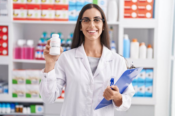 Die junge brünette Frau, die in der Apotheke arbeitet, hält Tabletten lächelnd mit einem fröhlichen und kühlen Lächeln im Gesicht. Zähne zeigen.  - Foto, Bild
