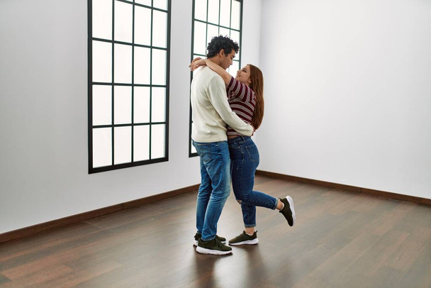 Νεαρό ζευγάρι ισπανόφωνων χαμογελά χαρούμενο και αγκαλιάζει στέκεται σε άδειο νέο σπίτι. - Φωτογραφία, εικόνα