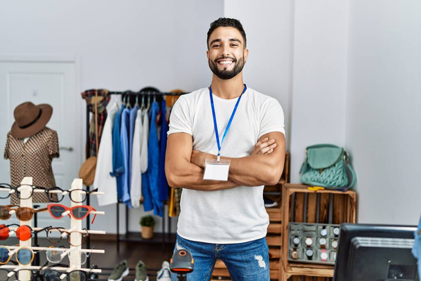 Young arab man shopkeeper smiling confident standing with arms crossed gesture at clothing store - Фото, изображение