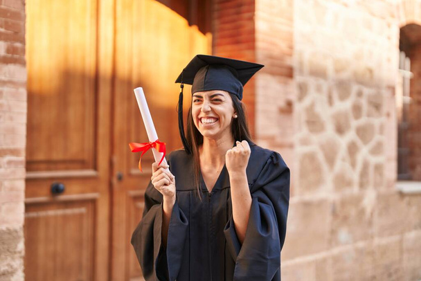 Young hispanic woman wearing graduated uniform holding diploma at university - Zdjęcie, obraz
