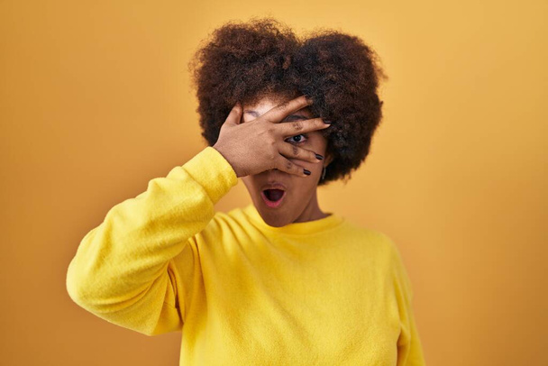 Молодая африканская американка, стоящая на жёлтом фоне, подглядывая в шокирующее лицо и глаза рукой, смотрит сквозь пальцы со смущенным выражением лица.  - Фото, изображение