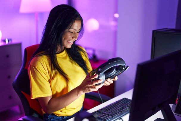 Young beautiful latin woman streamer smiling confident holding headphones at gaming room - Fotó, kép