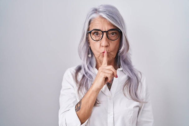 Donna di mezza età con tatuaggi che indossano occhiali in piedi su sfondo bianco chiedendo di essere tranquillo con il dito sulle labbra. silenzio e concetto segreto.  - Foto, immagini