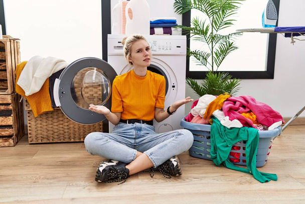 Junge blonde Frau beim Wäschewaschen sitzt ratlos und mit offenen Armen vor der Waschmaschine, keine Ahnung.  - Foto, Bild