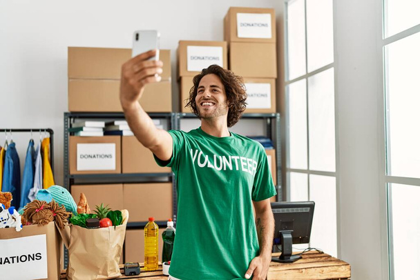 Joven voluntario hispano sonriendo feliz hacer selfie por el teléfono inteligente en el centro de caridad. - Foto, imagen