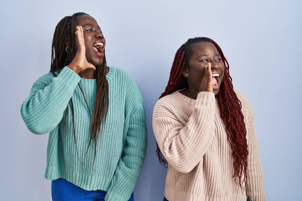 Zwei Afrikanerinnen stehen vor blauem Hintergrund und schreien laut Seite an Seite mit der Hand auf den Mund. Kommunikationskonzept.  - Foto, Bild