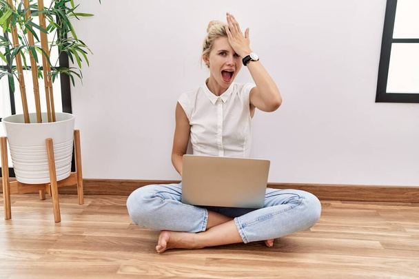 Young blonde woman using computer laptop sitting on the floor at the living room surprised with hand on head for mistake, remember error. forgot, bad memory concept.  - 写真・画像