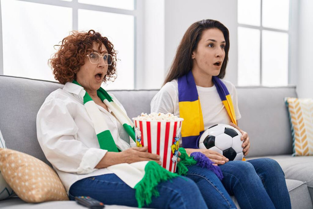 Hispanic mother and daughter watching football supporting team scared and amazed with open mouth for surprise, disbelief face  - Foto, afbeelding
