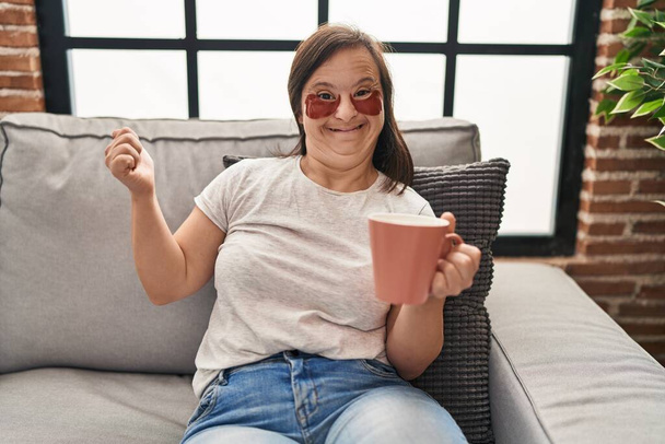 Chica hispana con síndrome de Down sentada en el sofá tomando un café en casa gritando orgullosa, celebrando la victoria y el éxito muy emocionada con el brazo levantado  - Foto, Imagen