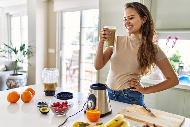 Молода красива іспаномовна жінка посміхається впевнено тримає склянку овочевої смузі на кухні
 - Фото, зображення