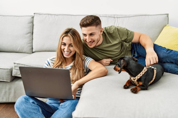 Młoda latynoska para uśmiecha się szczęśliwa korzystając z laptopa siedzącego na kanapie z psem w domu. - Zdjęcie, obraz