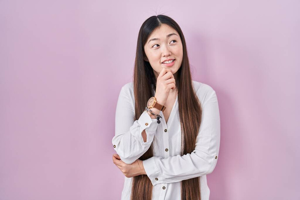 Chińska młoda kobieta stojąca na różowym tle z ręką na podbródku myśląca o pytaniu, przenikliwym wyrazie twarzy. Uśmiechnięta i troskliwa twarz. koncepcja wątpliwości.  - Zdjęcie, obraz