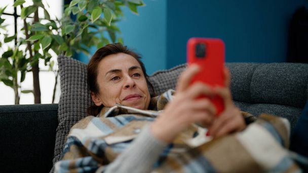 Střední věk hispánská žena pomocí smartphone ležící na pohovce doma - Fotografie, Obrázek