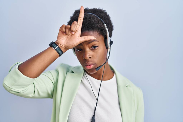 Mujer afroamericana usando auriculares de agente de centro de llamadas burlándose de personas con dedos en la frente haciendo gesto perdedor burlándose e insultando.  - Foto, Imagen