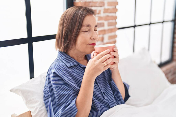 Középkorú nő iszik csésze kávét ül az ágyon a hálószobában - Fotó, kép