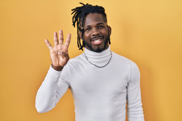 Afrikanischer Mann mit Dreadlocks trägt Rollkragenpullover über gelbem Hintergrund und zeigt mit Finger Nummer vier nach oben, während er selbstbewusst und glücklich lächelt.  - Foto, Bild