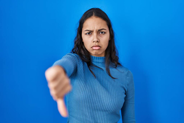 Młoda Brazylijka stojąca nad błękitnym, odizolowanym tłem, wyglądająca na nieszczęśliwą i wściekłą, wykazująca odrzucenie i negatyw gestem opuszczenia kciuków. złe wyrażenie.  - Zdjęcie, obraz