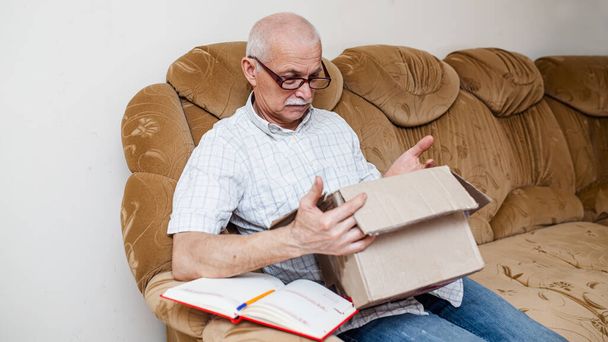 Hymyilevä iäkäs mies sai postipaketin kotona. Yllättyneellä isoisällä on avoin pahvilaatikko. Vanhuspäivystyksen tilaus - Valokuva, kuva