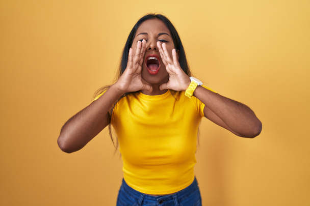 Giovane donna indiana in piedi su sfondo giallo gridando arrabbiato ad alta voce con le mani sulla bocca  - Foto, immagini