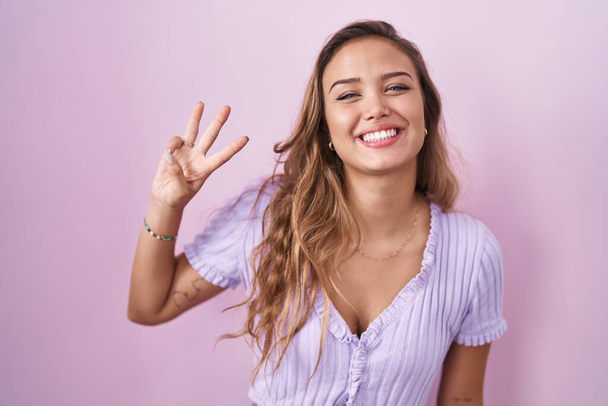 Mujer hispana joven de pie sobre fondo rosa mostrando y señalando hacia arriba con los dedos número tres mientras sonríe confiado y feliz.  - Foto, Imagen