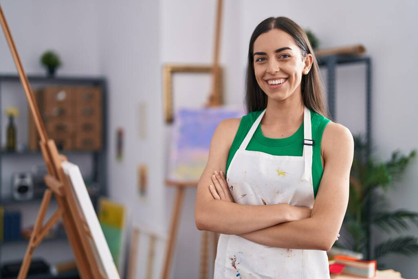 Молодая латиноамериканка-художница улыбается уверенно стоя со скрещенными руками в художественной студии - Фото, изображение