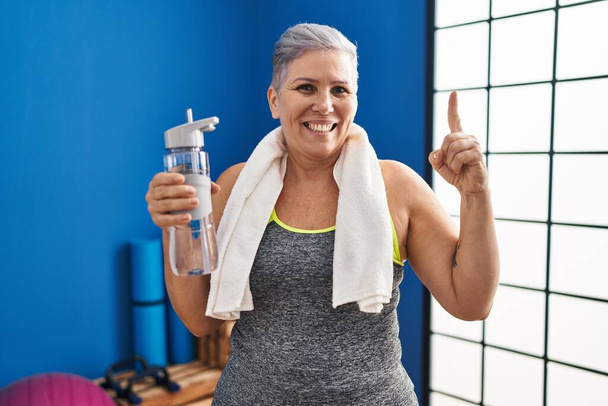 Middle age woman wearing sportswear and towel holding water smiling with an idea or question pointing finger with happy face, number one  - Zdjęcie, obraz