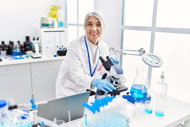 Mujer de pelo gris de mediana edad que usa uniforme científico usando microscopio en el laboratorio - Foto, imagen