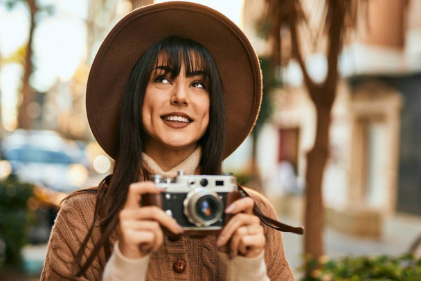 Brunette woman wearing winter hat smiling using vintage camera outdoors at the city - Foto, afbeelding