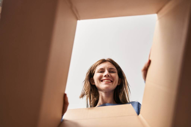 Güzel bir kadın, yüzünde mutlu ve havalı bir gülümsemeyle karton kutuyu açıyor. diş gösterme.  - Fotoğraf, Görsel