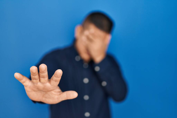 Латиноамериканец, стоящий на синем фоне, закрывая глаза руками и делая стоп-жест с грустным и испуганным выражением. смущенная и негативная концепция.  - Фото, изображение