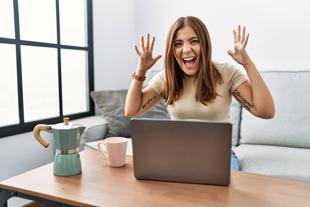 Young brunette woman using laptop at home drinking a cup of coffee celebrating crazy and amazed for success with arms raised and open eyes screaming excited. winner concept  - Foto, Bild