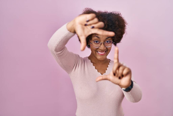 Молода афро-американська жінка, що стоїть над рожевим фоном, посміхається, роблячи рамку руками і пальцями з щасливим обличчям. Творчість і фотографія.  - Фото, зображення