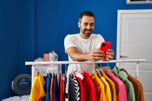 Fiatal spanyol férfi használ smartphone támaszkodó ruhák állvány mosókonyha - Fotó, kép