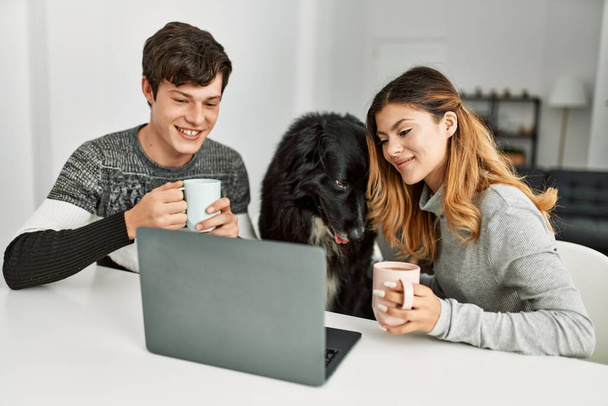 Jeune couple caucasien utilisant ordinateur portable et boire du café assis sur la table avec chien à la maison. - Photo, image