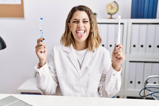 Jeune dentiste hispanique femme tenant brosse à dents électrique et brosse à dents à la clinique langue collante heureux avec une expression drôle.  - Photo, image