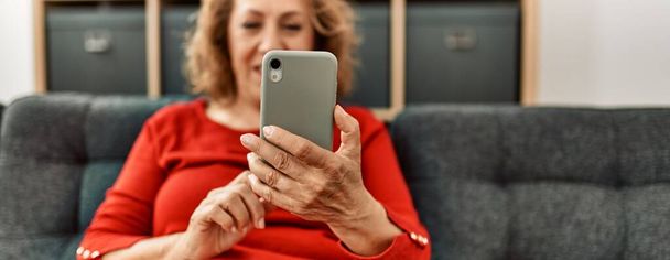 Middle age caucasian woman using smartphone sitting on the sofa at home. - Фото, изображение