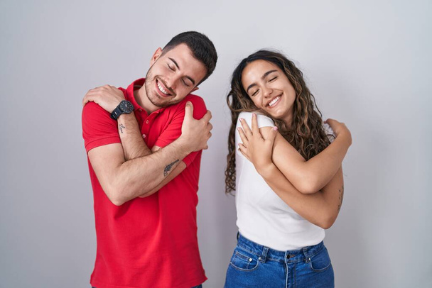 Young hispanic couple standing over isolated background hugging oneself happy and positive, smiling confident. self love and self care  - Foto, imagen