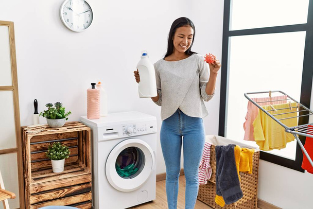 Joven latina sonriendo confiada sosteniendo detergente y juguete de virus en la lavandería - Foto, Imagen