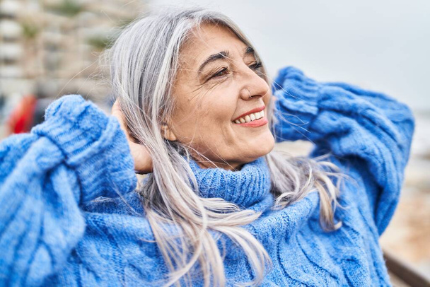 Šedovlasá žena středního věku, usmívající se sebevědomě uvolněná s rukama na hlavě u moře - Fotografie, Obrázek