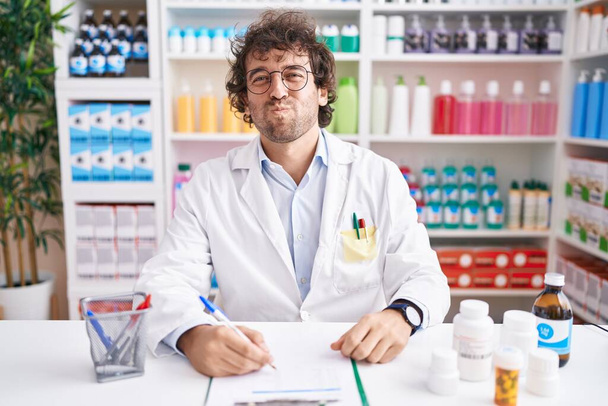 Hispanic young man working at pharmacy drugstore puffing cheeks with funny face. mouth inflated with air, crazy expression.  - Фото, изображение