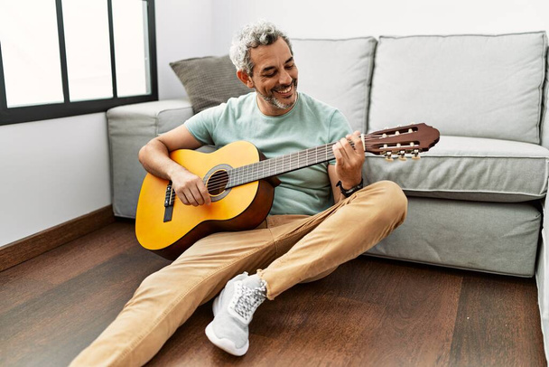 Średniowłosy mężczyzna w średnim wieku gra na gitarze klasycznej siedząc na podłodze w domu - Zdjęcie, obraz